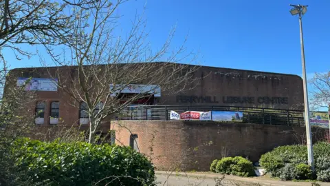 Ferryhill Sports and Education Centre is a large brown brick building with a ramp leading up to the reception. Bushes and trees are planted around the building.