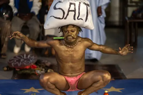 JEROME FAVRE/EPA Zobel Raoul in golden body paint squats as he grips a knife in his mouth and balances a bag of salt on his head. 