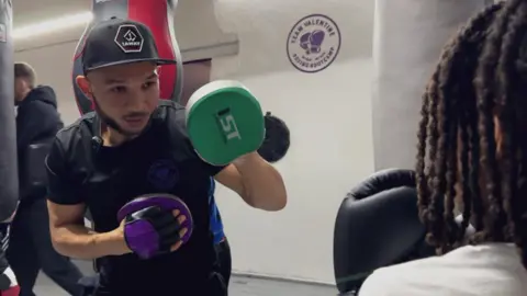 BBC Curtis is wearing a snapback, he has boxing gloves on, and is in his gym. He is wearing a black t-shirt and looks focused.