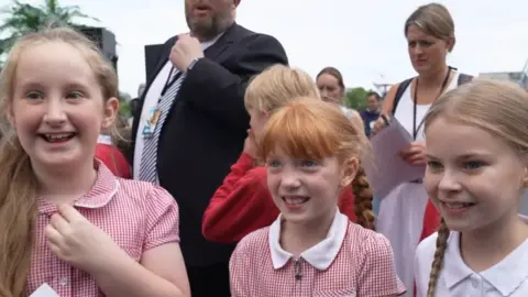 Three girls wearing red and white school uniforms. They are standing next to each other and looking to the side of the camera. They are all smiling. 