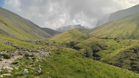 A general view of one of the paths up Scafell Pike. The rocky path is surrounded by grassy hills that are uneven and lead up to a tall mountain in the distance.