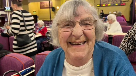 John Devine/BBC Jill Martin is an older woman with short, grey hair. She is smiling at the camera and is wearing spectacles and a blue cardigan. Some people sitting on chairs and walking around a bingo hall can be seen in the background