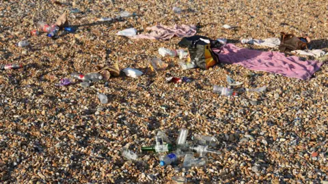 Eddie Mitchell Glass bottles, bags and towels discarded on a shingle beach.