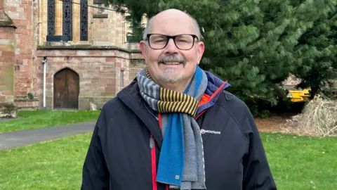 A man, with grey hair at the sides of his head and a grey goatee-style beard is standing in a churchyard. He is wearing dark-rimmed glasses, a colourful scarf and a dark fleece-type jacket.