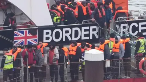 Refugees disembark from a Border Force boat. They are wearing orange life vests and are accompanied by officials.