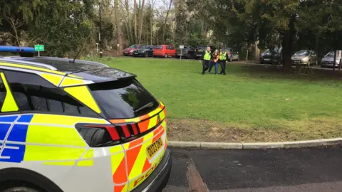 JOANNA TAYLOR/BBC The back of a police car with neon stripes is in the foreground. In the background, two police officers wearing hi-vis jackets stand on either side of Hazel Andrews-Oxlade who has her hands clasped together. They are walking towards the camera on grass. There is a car park with more vehicles in the background. 