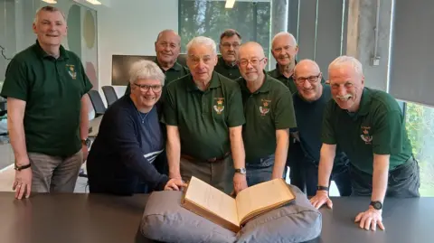 Sid Patterson, a trustee of the DLI Friends, centre, with Carolyn Ball, county archivist and manager of The Story, left, and Ian Brown, Chair of the DLI Friends, right, and other members of the Friends. They are smiling as the ledger rests on a big grey cushion in front of them. Sid Patterson has short white and is wearing a green t-shirt and grey trousers. Carolyn Ball has short white hair and blue long-sleeved top. She wears glasses and a necklace. Ian Brown has glasses and is wearing a blue top.