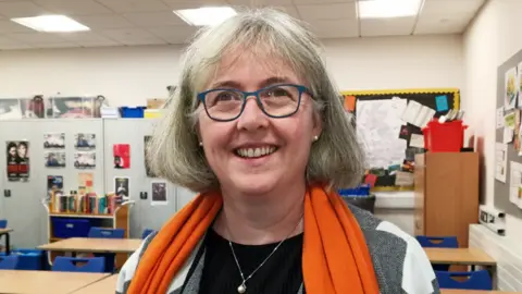 Victoria Rowe Victoria Rowe is a white woman in middle age. She has grey bobbed hair with a fringe. She wears a black top with a grey and white jacket and an orange scarf draped around her neck, as well as a silver necklace with a pearl pendant. She has blue-grey framed classes and is smiling. She is standing in a classroom with a few rows of desks and chairs behind her, cupboards with lots of pictures stuck to them and two pinboards on the walls.