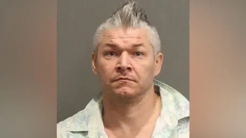 MNPD Joseph Rainey, who has grey, spikey hair and is wearing a pastel-coloured, open-collar shirt, in a police mugshot.