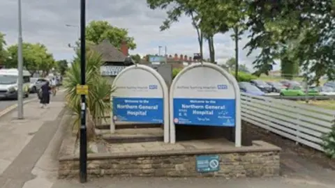 Google Blue and white NHS entrance signs at the Northern General Hospital in Sheffield with a wall in front, white fence to the right, black lamp-post to the left, woman in black walking away from the picture down the pavement on the right