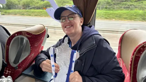 A person with a dark coat on wearing a hat. They are sat in a coach smiling holding a tinfoil FA Cup.