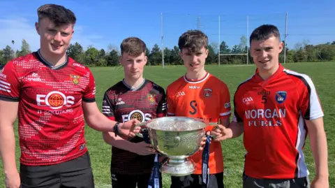 Four young men are pictured standing on a gaelic football pitch, each holding a large silver sports cup. 