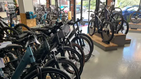 Într-un magazin sunt aliniate rânduri de biciclete electrice, cu anvelopele orientate spre exterior, în aceeași direcție. Trei biciclete electrice sunt montate separat pe scaune de lemn, în dreapta.