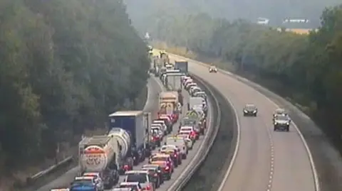 A motorway camera image of a motorway - there are two lanes of traffic queues on the left and a relatively empty carriageway - with just for cars visible - on the right. 