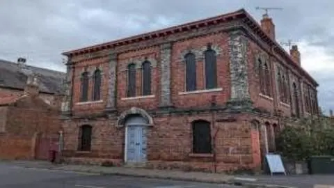 Exterior shot of the front of the red-bricked building, which has arched windows, a door and stone columns 