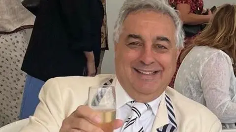 A middle-aged man with grey hair and wearing a cream suit, raises a glass of champagne to the camera