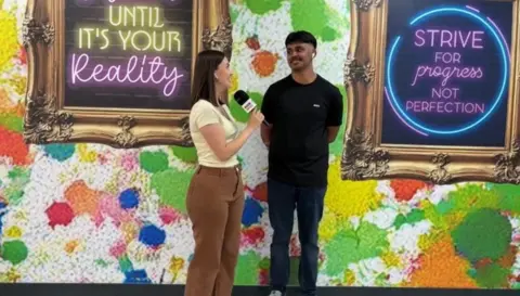Civ smiles as he speaks to a BBC reporter on A-Level results day. He is wearing a black t-shirt and blue jeans. The reporter is wearing brown trousers and a white top. 