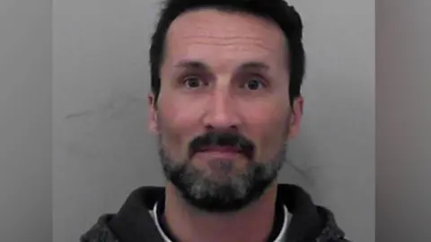 A police custody mugshot of Mark Acklom. He has a neat, dark beard with flecks of grey in it, and short dark hair. He is wearing a dark hoodie