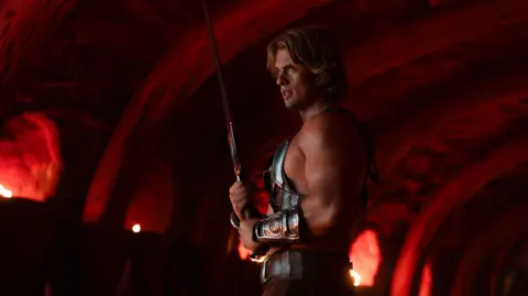 PA Media Nicholas Galitzine as He-Man. He is wearing body armour, shorts and is holding a large sword in a dark room. 
