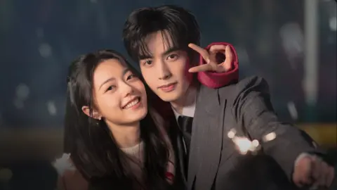 An attractive Chinese couple smile for the camera. The woman has long hair and has her arm around the man's shoulders, doing a peace sign, while the man who is dressed in a suit holds a lit sparkler towards the camera