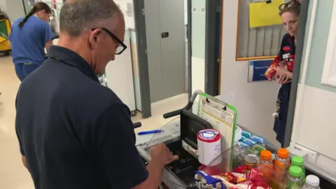 A man facing away from the camera. He is wearing a navy blue top. He is standing behind a trolly with crisps and drinks on and a small money tin. He is stood in a corridor. There are nurses around. 