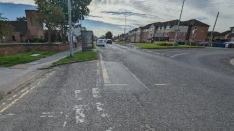 Stretch of worn road with edge of roundabout to the right of the shot and houses in the background.