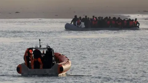 An orange inflatable French rescue vessel heading towards a large group of migrants on an inflatable dingy just off a sandy beach