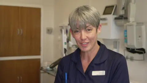 A midwife wearing blue scrubs looking at the camera in a hospital setting. She has short grey hair.