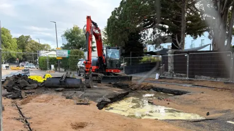 An area of road is cordoned off by construction fencing as a digger digs up the tarmac, surrounded by dirty water.