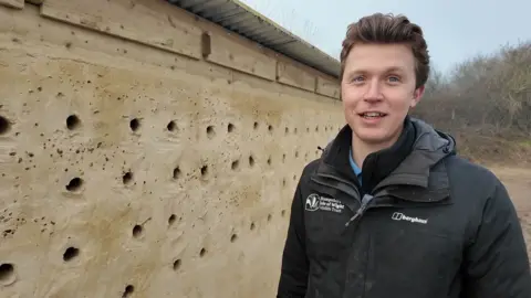 A man in a black jacket with dark brown hair stands in front of a sandy coloured low long building with scores of nest holes along the side