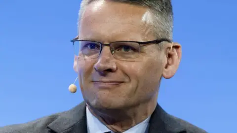 Reuters A close-up of John Pettigrew, who wears a suit and glasses and a small beige microphone that is hooked from his left ear, around the back of his head to stick out subtly to the right of his mouth