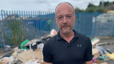 BBC Dave Hughes is standing in front of the rubbish heap which has blue railings to one side. He has short grey hair and a beard and is wearing a black polo shirt.