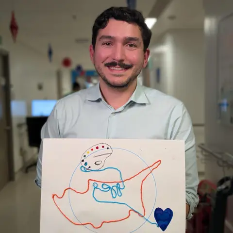 Dr Umur Guven, a man with dark hair and a moustache, wearing a shirt and holding up his artwork