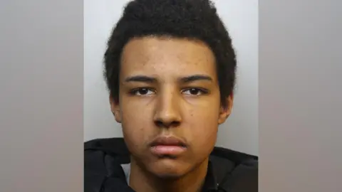 A mugshot of Romeo Dumbi. He has short black hair and some stubble on his upper lip. He is pictured looking directly at the camera with a blank expression.