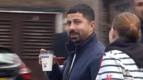 A man with short black hair and facial hair stares directly at the camera, mid-conversation with the woman next to him. He is wearing a navy zip-up hoodie and holding a coffee in a paper cup.