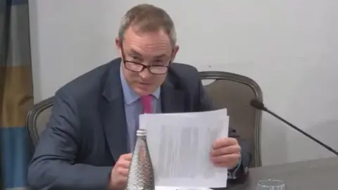 A still image of a livestream. Rob Sadler is speaking at the inquiry, holding papers in his hands. He has short grey hair and is wearing dark glasses, a navy suit, blue shirt and pink tie.