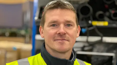 A man with short brown hair, a yellow reflective top and safety glasses on top of his head in a blurred room