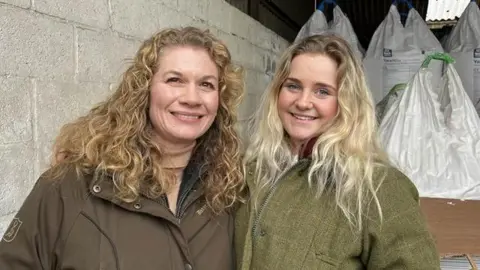 Phil Harrison/BBC A blonde woman with curly hair in a brown coat smiling and standing next to a younger blonde woman in a green tweed coat.