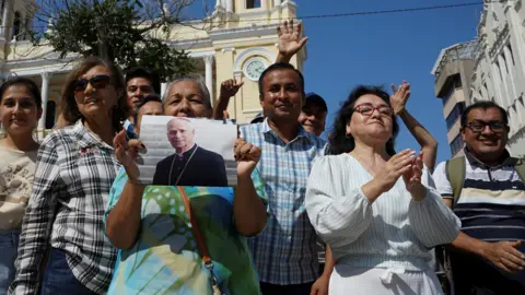 Reuters A group of people stand smiling in front of the cathedral in Chicalyo, Peru. One holds a picture of Pope Leo XIV.