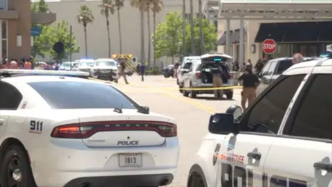 A close up picture of two police cars and other police cars in the distance and yellow police tape. Theer are palm trees and officers milling around the cars. The picture is near to the scene of a shooting at the Mall of Louisiana.