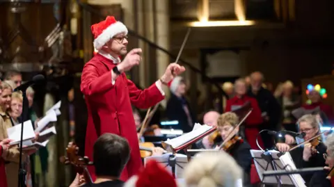Bethany Clarke A man wearing a santa hat and wearing a red robe conducts an orchestra in an old church building.