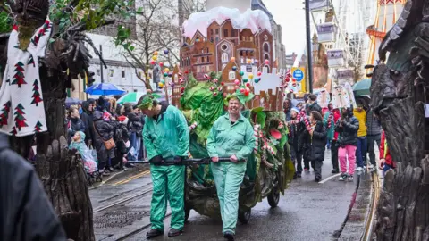 Manchester City Council A man and woman in full-body green suits drag a large prop of a multi-tiered gingerbread house. Children in the background wave red and white sticks and placards of gingerbread drawings. Spectators line the street on both sides.