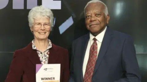 Hilda Moore holding her award smiling and standing to the left of Sir Trevor McDonald