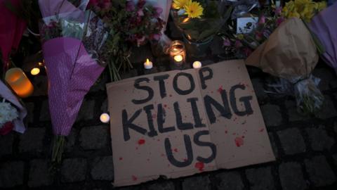 A cardboard sign saying "STOP KILLING US" is seen at a memorial site, among candles and flowers, in Clapham Common Bandstand, following the kidnap and murder of Sarah Everard