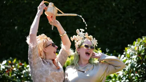 A woman is holding a wicker watering can over her friend's head. Her friend is wearing a fascinator with a floral theme. 