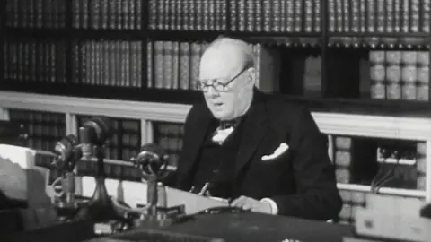 A black and white photo of Winston Churchill wearing glasses, sat behind a desk with microphones