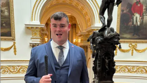 Brandon Hulcoop Brandon Hulcoop wears a blue blazer, waistoat and tie with a white shirt. Seen in Buckingham Palace, he smiles holding a stick.