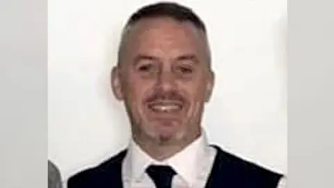 Phillip Thompson with very short dark hair, wearing a white shirt, dark tie and dark waistcoat, smiling to camera