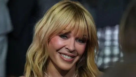 A close-up portrait of singer Kylie Minogue smiling. She has blonde, shoulder-length hair with a full fringe. She is wearing a pearl necklace. The background is blurred with dark tones and a grid pattern.
