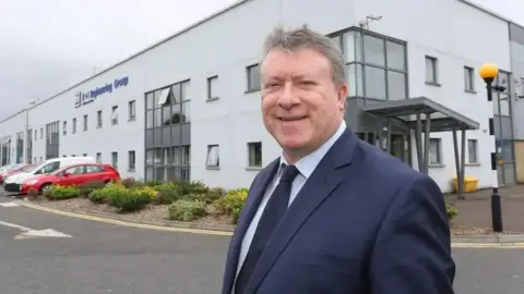 Philip O'Doherty has short dark grey hair and is wearing a navy suit and tie and is standing in front of an office building, which is modern and painted white with several grey-framed windows. Cars are parked to the left of the image.